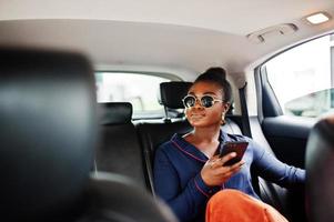 una rica mujer africana de negocios con gafas de sol se sienta en un camión suv con asientos de cuero negro. teléfono móvil a la mano. foto