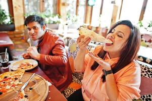 pareja de amigos asiáticos comiendo pizza durante la fiesta en la pizzería. gente india feliz divirtiéndose juntos, comiendo comida italiana y sentados en el sofá. foto