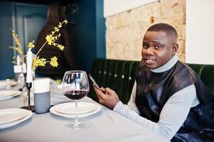 hombre africano con ropa tradicional negra sentado en el restaurante con una copa de vino tinto con el teléfono celular a mano. foto