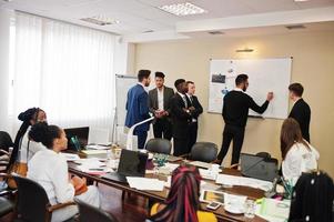 Multiracial business team addressing meeting around boardroom table and write plan on board. photo