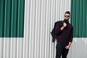 Fashion rich beard Arab man wear on turtle neck and sunglasses posed against green and white steel wall with jacket on hand. Stylish, succesful and fashionable arabian model guy. photo