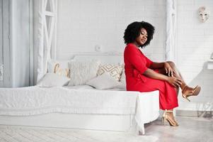 Fashion african american model in red beauty dress, sexy woman posing evening gown sitting at bed in white vintage room. photo