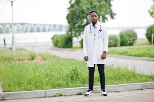 joven médico afroamericano de bata blanca con un estetoscopio posado al aire libre. foto