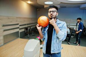 Stylish asian man in jeans jacket and glasses standing at bowling alley with ball at hand. photo