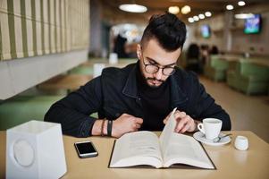 Arab man wear on black jeans jacket and eyeglasses sitting in cafe, read book and drink coffee. Stylish and fashionable arabian model guy. photo