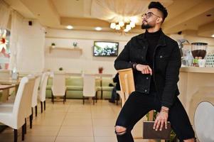 Arab man wear on black jeans jacket and eyeglasses in cafe drink coffee at bar with book. Stylish and fashionable arabian model guy. photo