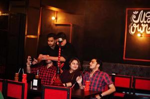 Group of indian friends having fun and rest at night club, drinking cocktails and smoke hookah, looking at mobile phones. photo