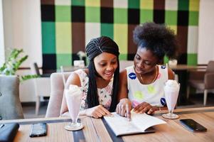 Two black african girlfriends at summer dresses drinking milkshake cocktails and looking in menu at cafe. photo