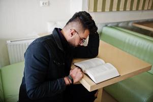 el hombre árabe usa una chaqueta de jeans negros y anteojos sentados en un café, lee un libro. chico modelo árabe elegante y de moda. foto