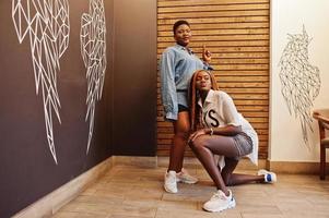 Two african womans in stylish casual clothes posing against wooden wall sat down and dance twerk. photo