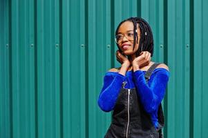 African woman in denim overalls skirt, eyeglasses posed against green steel wall. photo