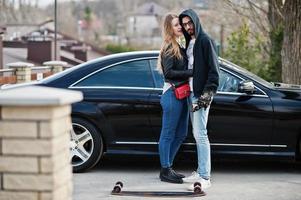 Cool pareja multirracial se abraza junto con longboard contra elegante coche deportivo negro. foto