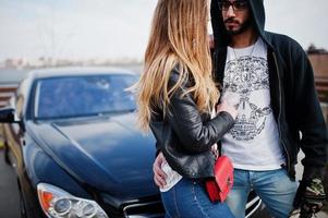 una pareja multirracial genial se abraza contra un elegante auto deportivo negro. foto