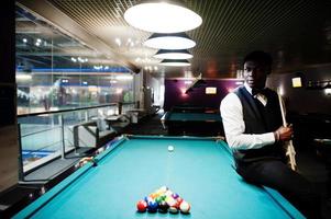 Young handsome african man wearing white shirt, black vest and bow tie play pool billiard. photo