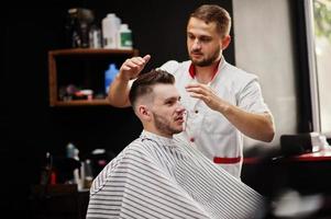 Young bearded man getting haircut by hairdresser while sitting in chair at barbershop. Barber soul. photo