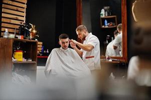 Young bearded man getting haircut by hairdresser while sitting in chair at barbershop. Barber soul. photo