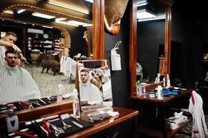 Young bearded man getting haircut by hairdresser while sitting in chair at barbershop. Barber soul. photo