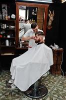 Young bearded man getting haircut by hairdresser while sitting in chair at barbershop. Barber soul. photo