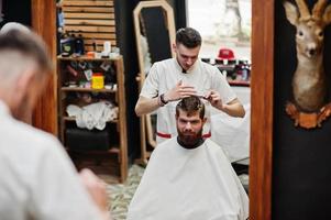 Young bearded man getting haircut by hairdresser while sitting in chair at barbershop. Barber soul. photo