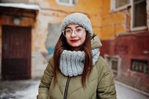 Portrait of brunette girl in gray scarf and hat, glasses at cold weather against orange wall of old house. photo