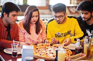 Group of asian friends eating pizza during party at pizzeria. Happy indian people having fun together, eating italian food and sitting on couch. photo