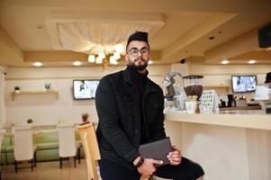 Arab man wear on black jeans jacket and eyeglasses in cafe drink coffee at bar with book. Stylish and fashionable arabian model guy. photo