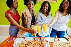 cuatro jóvenes africanas en un restaurante de pizza de colores brillantes tintineando jugos. foto