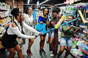 grupo de cinco mujeres africanas con trapeador, cepillo de baño y balde divirtiéndose en el departamento de artículos de limpieza del hogar en el supermercado. foto