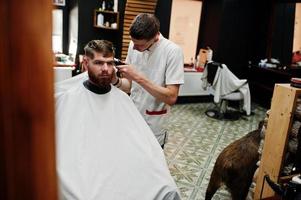 Young bearded man getting haircut by hairdresser while sitting in chair at barbershop. Barber soul. photo