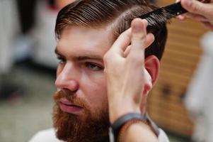 Young bearded man getting haircut by hairdresser while sitting in chair at barbershop. Barber soul. photo