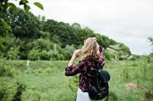 retrato de una atractiva chica rubia con camisa de tartán con mochila caminando por el campo. foto