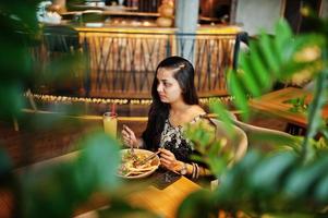 linda chica india vestida con sari negro posó en el restaurante, sentada a la mesa con jugo y ensalada. foto