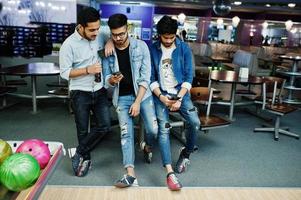 tres amigos del sur de asia con camisa de jeans en el club de bolos mirando algo en sus teléfonos móviles. foto