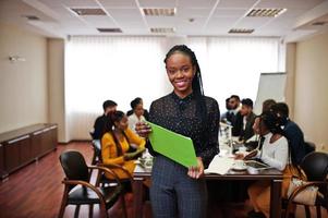 Face of handsome african business woman, holding clipboard on the background of business peoples multiracial team meeting, sitting in office table. photo
