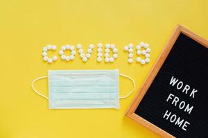 Flat lay of medicine pills shape as word covid 19 with mask and notice board with message work from home on yellow background, prevent the spread of pandemic Covid-19 and Coronavirus photo