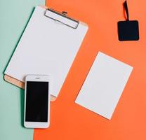 Creative flat lay of smartphone, blank clipboard and white card on minimal color background photo