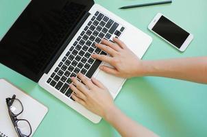 Hand using laptop on creative flat lay workspace desk with smartphone and stationery on green background photo