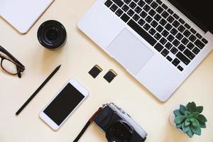 Desktop flat lay photo of workspace desk with laptop, camera, tablet, smartphone, lens and notebook on yellow modern background, mood board style