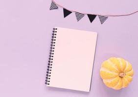 Flat lay of minimal workspace blank notebook with pumpkin and bunting flag on purple background, copy space photo