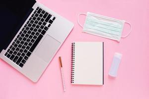 Flat lay of workspace desk with laptop, blank notebook, medical masks and alcohol gel on bright pink background, work from home and self quarantine concept, prevent the Covid-19 and Coronavirus