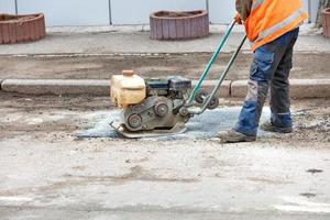 A road worker is patching up a pothole in an old road using an old petrol plate compactor. photo