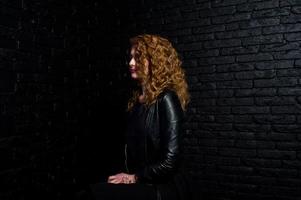 Curly hair girl at leather jacket on studio against black brick wall. photo