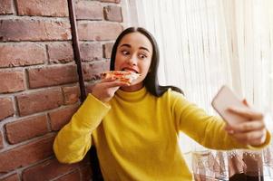 chica morena divertida en suéter amarillo comiendo pizza en el restaurante y haciendo selfie. foto