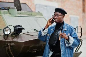 African american man in jeans jacket, beret and eyeglasses, with cigar posed against btr military armored vehicle, and speaking on mobile phone. photo