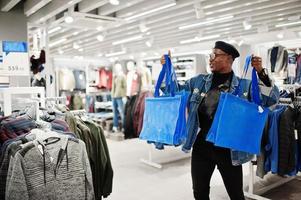 Stylish casual african american man at jeans jacket and black beret with fanny pack or waist bag holding blue shopping bags at clothes store. photo