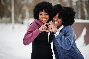 dos mujeres afroamericanas de cabello rizado usan suéteres posados en el día de invierno, muestran el corazón con los dedos. foto