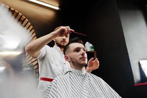 Young bearded man getting haircut by hairdresser while sitting in chair at barbershop. Barber soul. photo