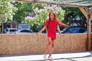 Cute and slim african american girl in red dress with dreadlocks in motion having fun on street. Stylish black model. photo