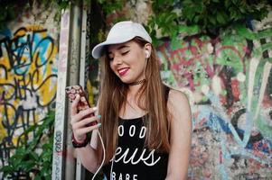 Stylish casual hipster girl in cap and jeans wear, listening music from headphones of mobile phone against large graffiti wall. photo