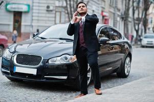 Stylish indian businessman in formal wear with mobile phone standing against black business car on street of city. Speak on cellphone and looking at his watches. photo